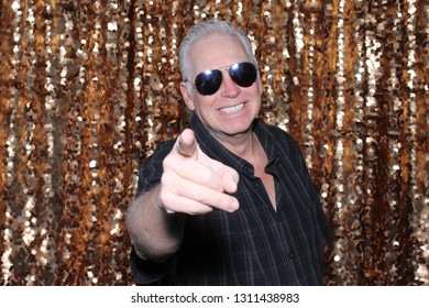 Man In A Photo Booth. A Caucasian Man Smiles And Has Fun Posing In A Photo Booth With A Gold Sequin Curtain. White Male In Sunglasses Points To The Camera And Smiles.
 