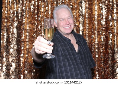 Man In A Photo Booth. A Caucasian Man Smiles And Has Fun Posing In A Photo Booth With A Gold Sequin Curtain. White Male Drinks And Toast A Champagne Glass Into The Camera And Smiles.
