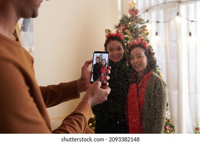 Man Photographing Wife And Mother In Law Under Falling Aftificial Snow At Christmas Tree At Home