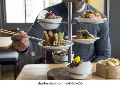 Man Picking Tofu Using Chopsticks From Tiered Server Filled With Vegetarian Dishes