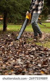 A man picks up fallen leaves using an electric leaf blower. Cordless Leaf Blowers. handheld, cordless, electric leaf blower in a garden, selective focus. Autumn, fall gardening works in a backyard