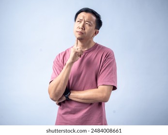 A man in a pink shirt looks thoughtfully upwards with his hand on his chin, reflecting a pensive and contemplative mood. He stands against a plain light blue background - Powered by Shutterstock