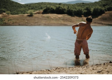 A man in pink shorts skipping stones in a peaceful lake surrounded by lush green hills under a clear blue sky. - Powered by Shutterstock