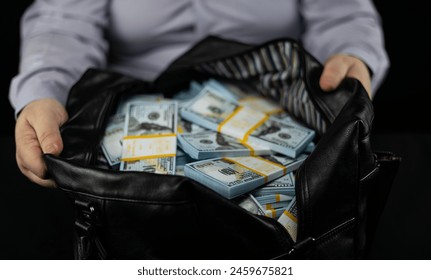 The man places a large bag on a black background and unzips it to reveal a large amount of dollars inside. Having unzipped the bag, the man looks inside and sees a million dollars in bundles. - Powered by Shutterstock