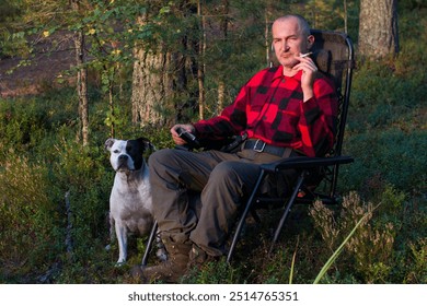 a man in a plaid shirt with a cigarette in his hand is sitting in an armchair in the woods. Wild outdoor recreation - Powered by Shutterstock