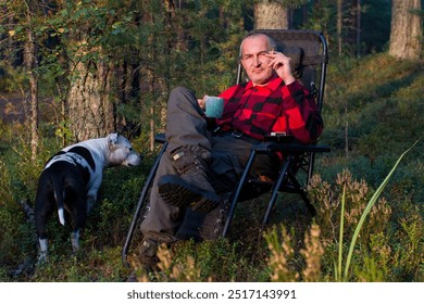 a man in a plaid shirt with a cigarette in his hand is sitting in an armchair in the woods. Wild outdoor recreation - Powered by Shutterstock