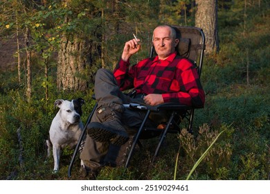 a man in a plaid shirt with a cigarette in his hand is sitting in an armchair in the woods. Wild outdoor recreation - Powered by Shutterstock