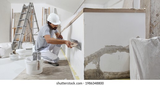 man plasterer construction worker at work, takes plaster from bucket and puts it on trowel to plastering the wall, wears helmet inside the building site of a house