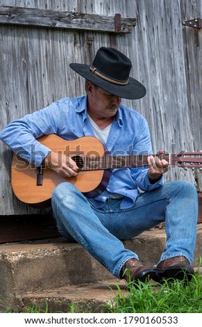 Man playing acoustic guitar alone outdoors sitting in front of old gray country wood barn wearing cowboy hat, rural country, farm, ranch, vintage, independent, musician, music, musical instrument, 