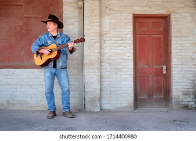 Man playing acoustic guitar outside in front of brick wall wearing cowboy hat and jeans in the city, on dirty porch, Spanish classical guitar