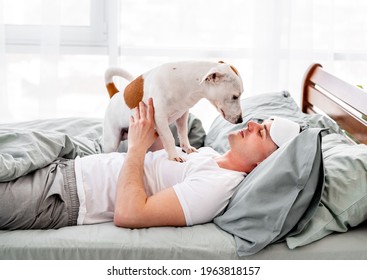 Man Playing With Dog In The Bed In The Morning Time. Young Guy Wakes Up And Doggy Pet Standing On Him And Licks His Face. Concept Of Friendship Beetween Human And Animal