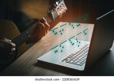 A man playing guitar in living room  while using laptop open chord on website. young guys learning music with a guitar chord table.