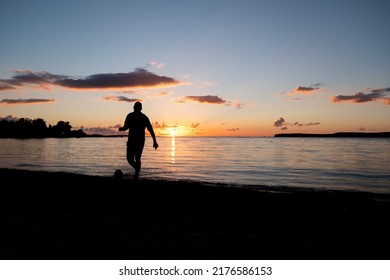 Man Playing Soccer On A Beach At Sunset. Dark Evening View. Football Player Silhouette On A Sandy Beach Late Evening.