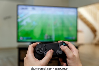 Man playing video game. Hands holding console controller. Football or soccer game on the television. Widescreen tv stands on commode. - Shutterstock ID 583000624