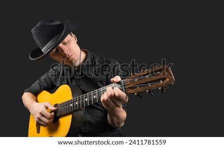 Man plays an acoustic guitar on a dark isolated background. A musician in a cowboy hat clamps the frets of a guitar on the neck. Musical instrument. Place for text. Copy space.