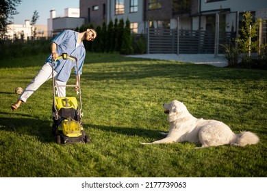 Man plays with a dog while moving lawn with a lawn mower at backyard of country house. Husband spending summer time with pet happily
