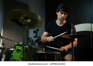 A man plays drums in a home studio at night. He is seated behind a drum set and uses drumsticks to hit the drums and cymbals. He is wearing a black t-shirt and black shorts. The room is dimly lit - Powered by Shutterstock