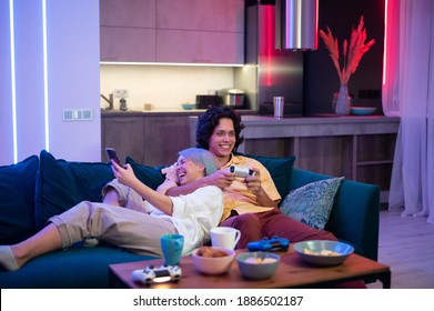 Man Plays On Gaming Console Using Joystick While Woman Using Smart Phone With Gaming App. Millenial Couple Lay On A Couch At Home And Play Video Games.