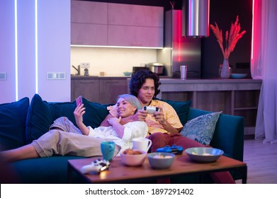 Man Plays On Gaming Console Using Joystick While Woman Using Smart Phone With Gaming App. Millenial Couple Lay On A Couch At Home And Play Video Games.