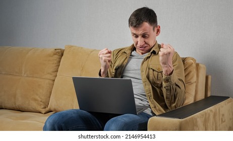 Man plays online gambling games on computer sitting on soft couch near grey wall at home. Expressive player lost money screaming and hitting armrest - Shutterstock ID 2146954413