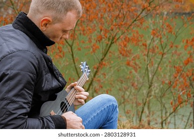 Man plays ukulele while spending time outdoors in autumnal natural setting. Musician holds small string instrument and performs music in relaxed atmosphere surrounded by greenery and open space