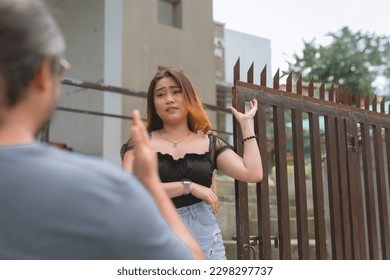 A man pleads to a younger lady in a black blouse standing beside the gate of her home. Not believing in his excuses and alibis. - Shutterstock ID 2298297737