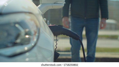 Man Plugging In Electric Car At Home At Electro Charging Station. Luxury White Electrical Car Recharging. Environmentally Conscious Man Charging His Electric Vehicle.