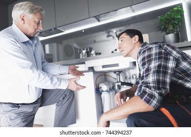 A Man Plumber Speaks With A Client Who Is Dissatisfied With The Work Done. The Old Man Communicates With The Plumber Who Sits Next To The Sink.