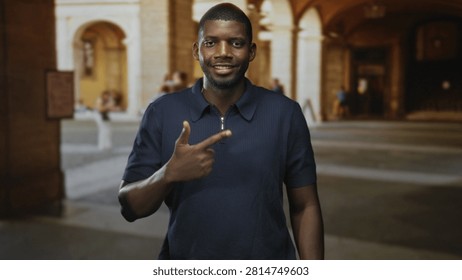 Man pointing finger to chest while smiling in a building arcade with stone arches, blurred passersby and tiled floor; confidence greeting.