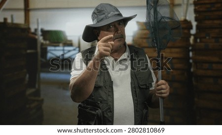 Man pointing finger while holding a fishing landing net, wearing a brim hat and fishing vest inside a dim warehouse stacked with crates; firm warning.