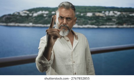 Man points finger at camera with raised hand on seaside railing, beard visible and stern face; anger warning.