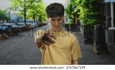Man points finger toward camera from outstretched arm on cobbled street in city wearing yellow t shirt; accusation.