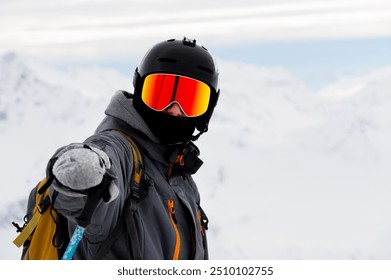 man points his hand at the camera in sunglasses with a ski helmet on his head and holds a stick in his hand against the background of white snow and mountains