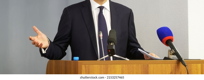 Man - politician, lawyer or official gesturing with his hands, holding documents and speaking from the podium in front of microphones. Speaker, orator, presenter or interviewee. No face. Web banner