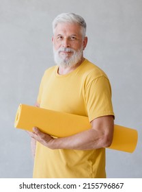 A Man Poses In A Sports Class, Holding A Yellow Yoga And Fitness Mat In His Hands. Senior Yoga At Home Or In The Gym For Training. A Fifty-year-old Man With Gray Hair Is Doing A Warm-up