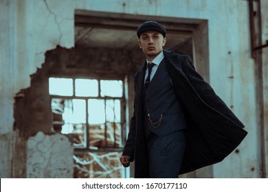 A man posing in the image of an English retro gangster of the 1920s dressed in a coat, suit and flat cap in Peaky blinders style.