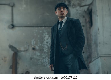 A man posing in the image of an English retro gangster of the 1920s dressed in a coat, suit and flat cap in Peaky blinders style.