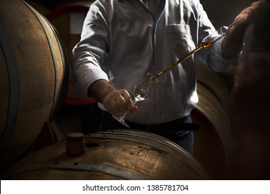 Man, pouring cognac from the barrel into glass in old rustic underground wine cellar with rows of big oak barrels. Famous wine and brandy industrial destination