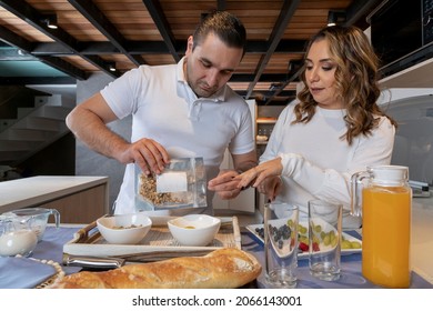 Man Pouring Granola Into A Bowl With Yogurt While Making Breakfast With His Wife