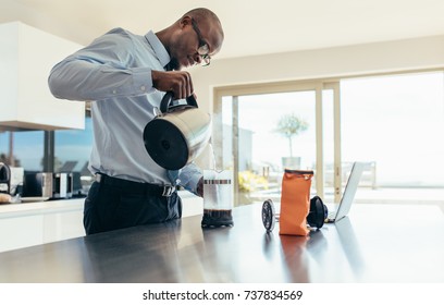 Man pouring hot water in coffee maker. Businessman preparing coffee on breakfast table with a laptop computer by the side.