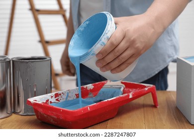 Man pouring light blue paint from bucket into tray at wooden table indoors, closeup
