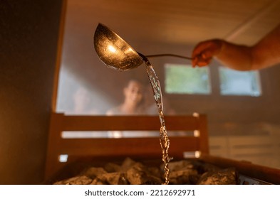 Man pouring water onto hot stone with metal spo in sauna room with a group of people. Steam an water on the stones, spa and wellness concept, relax in hot finnish sauna. Warm temperature bath therapy.