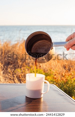 man pours freshly brewed coffee from an aluminum ladle into a mug on a tourist table, on the seashore. Travel and tourism.