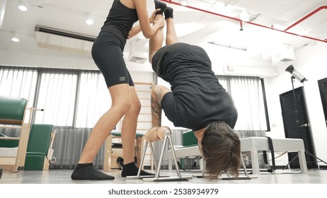 Man practicing assisted handstand on pilates reformer for core strength, balance, and upper body control at gym. Female trainer assist student handstand on reformer for improving balance. Habituate. - Powered by Shutterstock