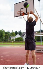 Man Practicing His Basketball Aiming For A Goal Tossing The Ball In The Air On An Outdoor All Weather Court Viewed From Behind