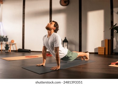 A man practicing yoga indoors, performing the upward-facing dog pose on a black yoga mat. The room has wooden floors and other yoga mats and props are visible in the background. - Powered by Shutterstock