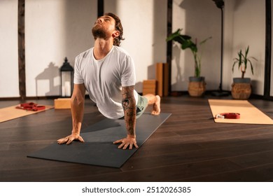 A man practicing yoga indoors, performing the upward-facing dog pose on a black yoga mat. The room has wooden floors and other yoga mats and props are visible in the background. - Powered by Shutterstock