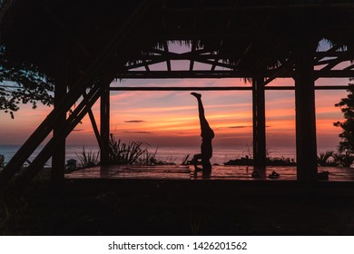 Man practising a headstand with sunset background. Yoga retreat with bamboo hut and red, orange sky. Handstand balance. Shot in Montanita beach, Ecuador.