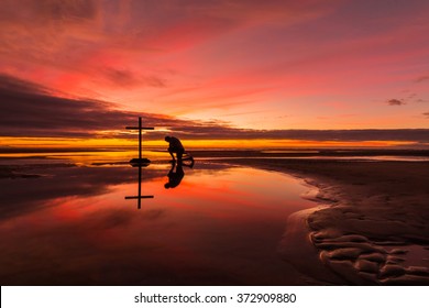 Man praying at a cross on a beach with a wonderful sunset sky behind him.