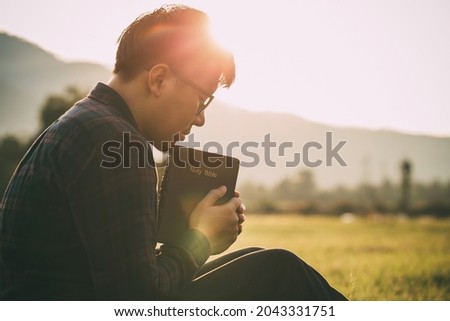 man praying on the holy bible in a field during beautiful sunset.male sitting with closed eyes with the Bible in his hands, Concept for faith, spirituality, and religion.

